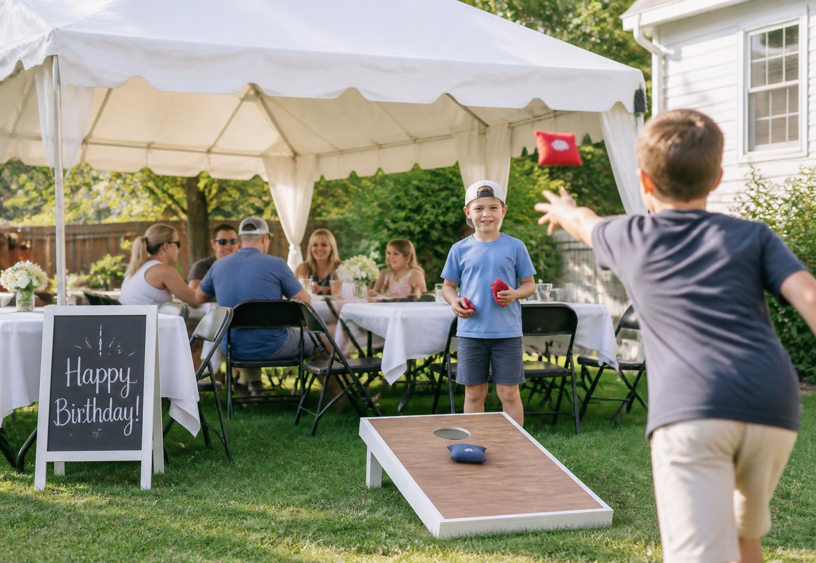 Family birthday party in a backyard with a white canopy tent, tables, chairs, and kids playing cornhole